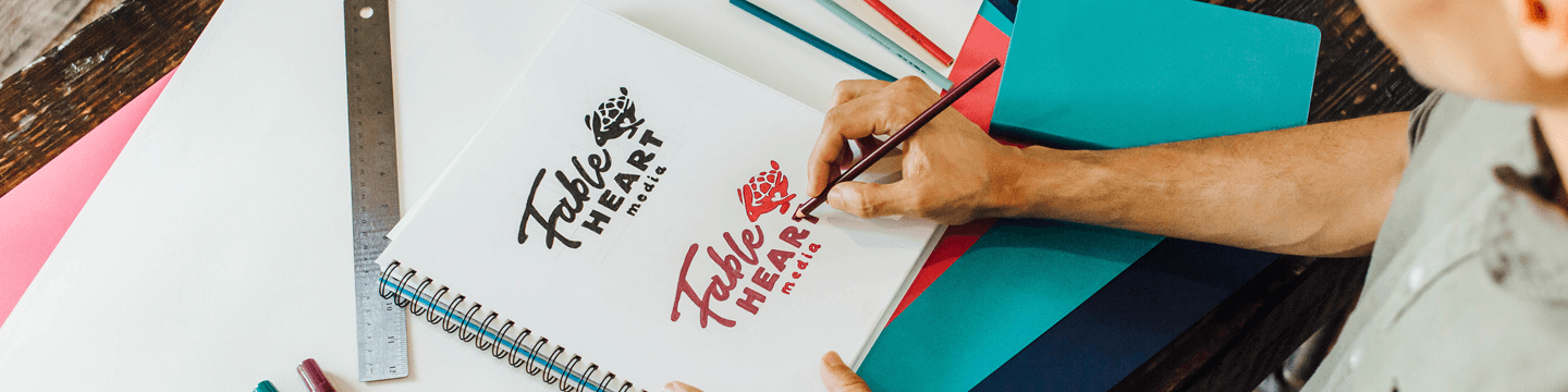 Someone drawing the Fable Heart Media logo surrounded by multi-colored paper.